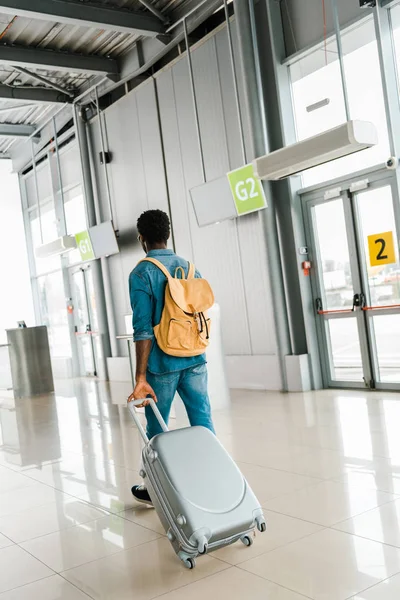 Vista trasera del hombre afroamericano caminando con maleta y mochila en el aeropuerto - foto de stock