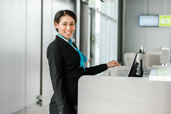 Smiling african american airport staff in uniform standing at check in desk and looking at camera — Stock Photo