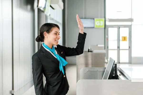 Улыбающийся африканский американский персонал аэропорта в форме стоя у стойки регистрации и размахивая рукой — стоковое фото