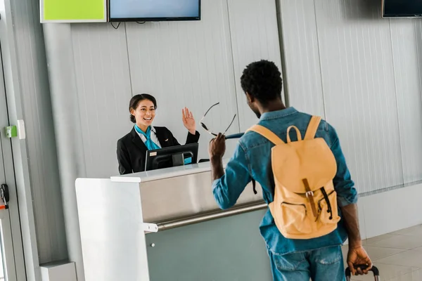 Smiling african american airport worker waving hand to tourist with backpack — Stock Photo