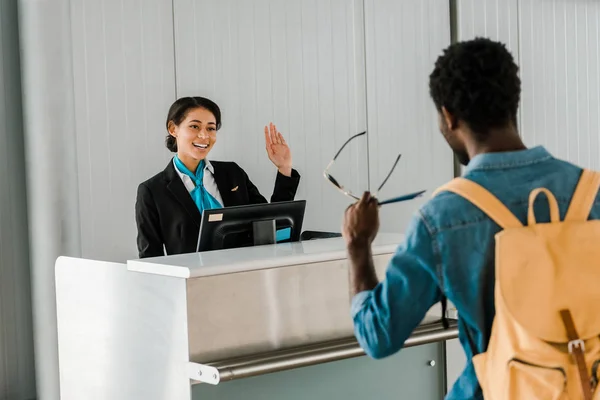 Selective focus of smiling african american airport worker waving hand to tourist with backpack — Stock Photo
