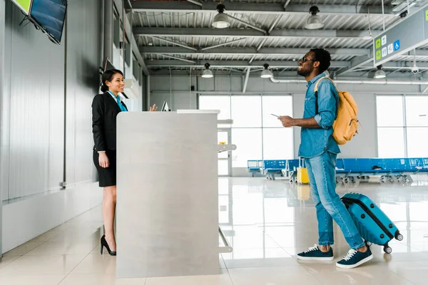 Side view of smiling african american airport worker and tourist with backpack and suitcase — Stock Photo