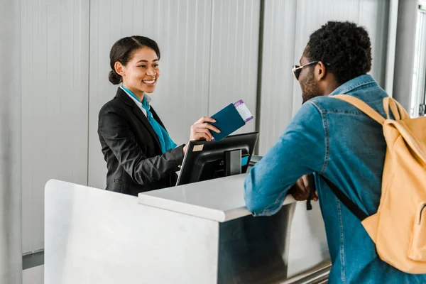 Sonriente afroamericano trabajador del aeropuerto dando pasaporte y billete de avión a turista con mochila - foto de stock