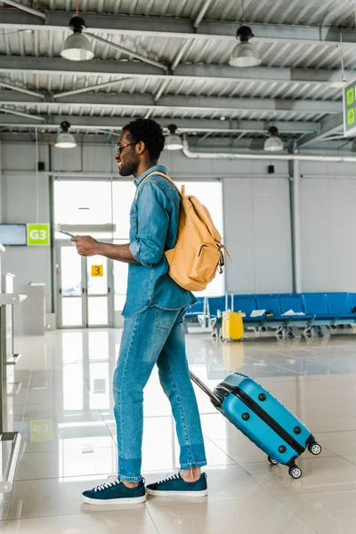 Fröhlicher afrikanisch-amerikanischer Mann im Jeans-Outfit mit Koffer und Rucksack im Flughafen — Stockfoto