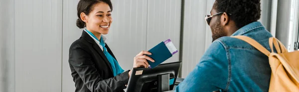 Panoramic shot of smiling african american airport worker giving passport and air ticket to tourist with backpack — Stock Photo
