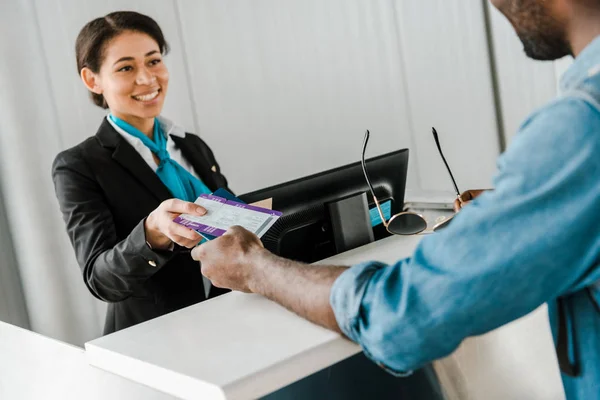 Alegre afroamericano trabajador del aeropuerto dando pasaporte y billete de avión al turista - foto de stock