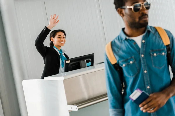 Sonriente afroamericano trabajador del aeropuerto saludando mano cerca de pasajero en gafas de sol con pasaporte y billete de avión - foto de stock