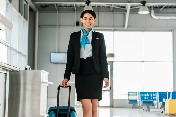 Sonriente atractiva azafata afroamericana caminando con la maleta en el aeropuerto - foto de stock