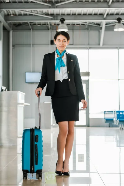Hermosa azafata afroamericana de pie con la maleta en el aeropuerto - foto de stock