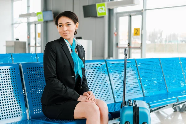 Attraktive afrikanisch-amerikanische Stewardess sitzt mit Koffer in der Abflughalle — Stockfoto