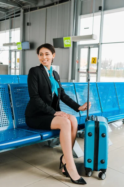 Sonriente azafata afroamericana sentada con la maleta en la sala de salida - foto de stock