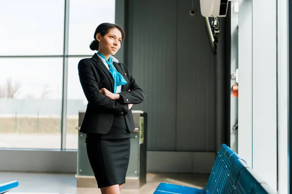 Hostess afroamericana in piedi con le braccia incrociate in aeroporto e guardando la finestra — Foto stock