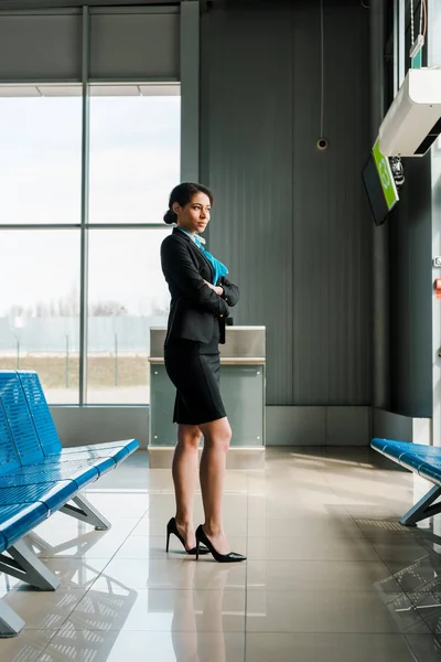 Hermosa azafata afroamericana de pie con los brazos cruzados en el aeropuerto y mirando hacia otro lado - foto de stock