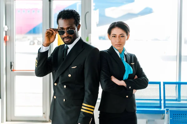 Seriöser afrikanisch-amerikanischer Pilot mit Sonnenbrille und Stewardess mit verschränkten Armen, die zusammen auf dem Flughafen stehen — Stockfoto