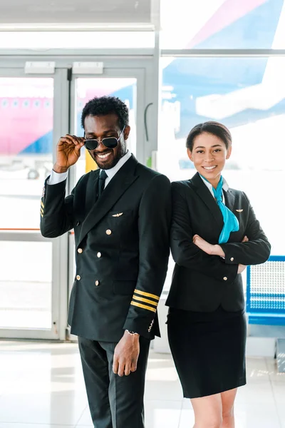 Piloto afroamericano en gafas de sol y azafata con brazos cruzados posando juntos en el aeropuerto - foto de stock
