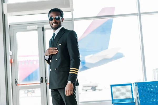 Улыбающийся африканский американский пилот, стоящий в зале вылета в аэропорту — стоковое фото