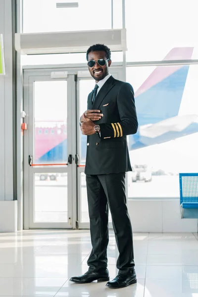 Smiling african american pilot in sunglasses showing thumb up in departure lounge in airport — Stock Photo