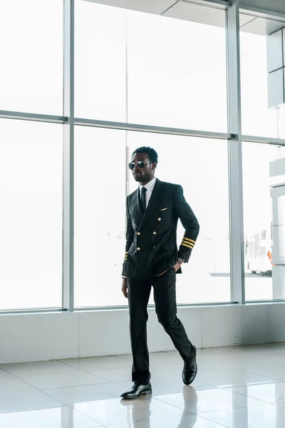 Piloto africano americano confiado en gafas de sol caminando en el aeropuerto con la mano en el bolsillo - foto de stock
