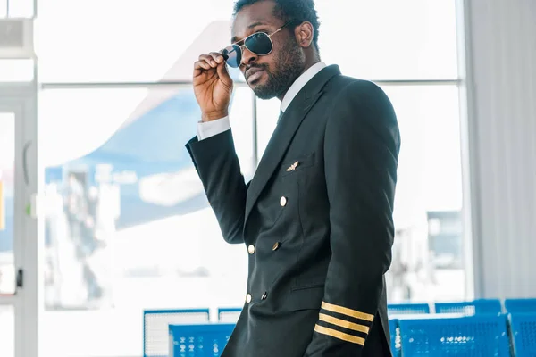 Elegante piloto afroamericano confiado en gafas de sol en la sala de salida en el aeropuerto - foto de stock