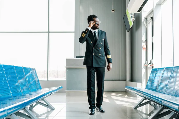 Piloto africano americano confiado en gafas de sol caminando en la sala de salida en el aeropuerto y mirando a la ventana - foto de stock
