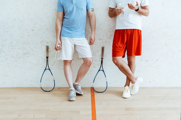 Cropped view of two squash players in shorts standing with crossed legs in four-walled court — Stock Photo