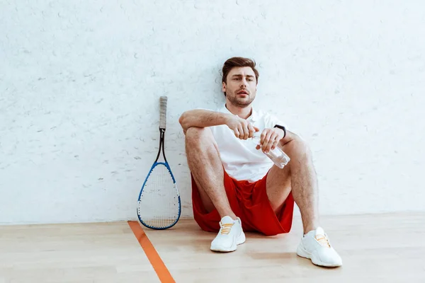 Müder Squash-Spieler sitzt auf dem Boden und öffnet Wasserflasche — Stockfoto