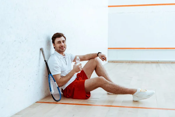 Squash jugador sentado en el suelo y sosteniendo la botella de agua - foto de stock