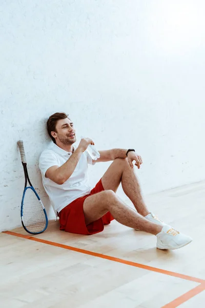 Jugador de squash en pantalones cortos rojos sentado en el suelo y agua potable - foto de stock