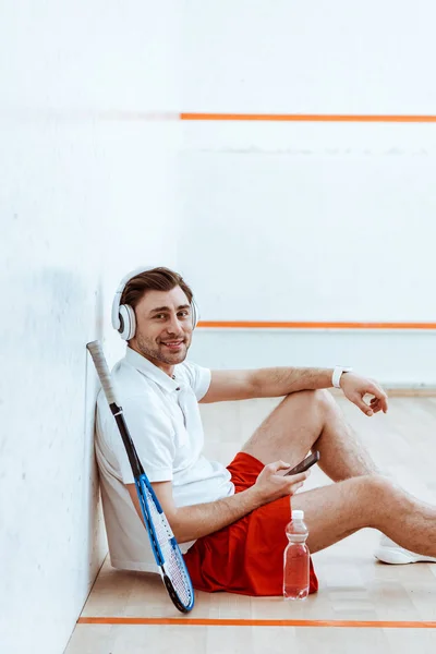 Sonriente reproductor de squash escuchar música en los auriculares y el uso de teléfonos inteligentes - foto de stock