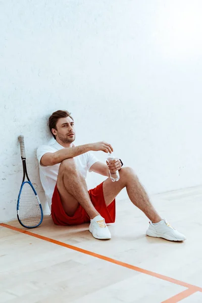 Squash jugador sentado en el suelo y sosteniendo la botella de agua — Stock Photo