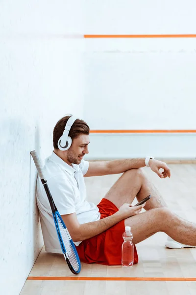 Sonriente reproductor de squash escuchar música en los auriculares y el uso de teléfonos inteligentes - foto de stock