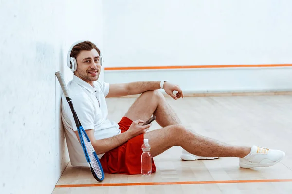 Squash player sorridente seduto sul pavimento e ascoltare musica in cuffia — Foto stock