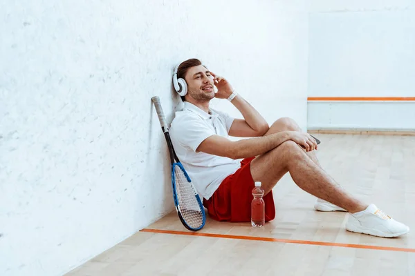 Reproductor de squash sonriente sentado en el suelo y escuchando música en los auriculares - foto de stock