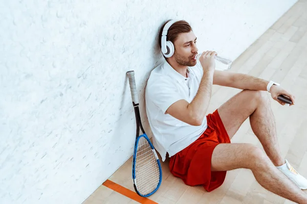 Vista de ángulo alto del reproductor de squash escuchando música en auriculares y agua potable - foto de stock