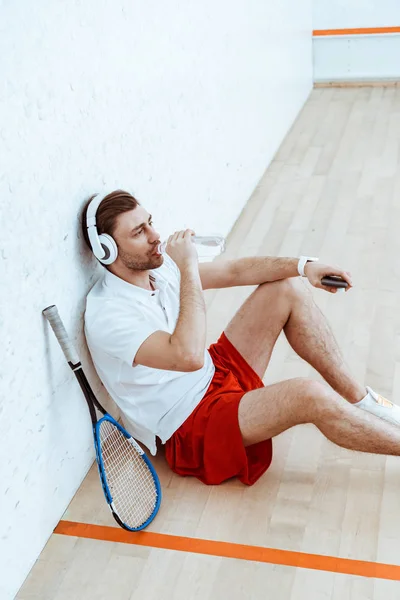 Vista de ángulo alto del reproductor de squash escuchando música en auriculares y agua potable - foto de stock