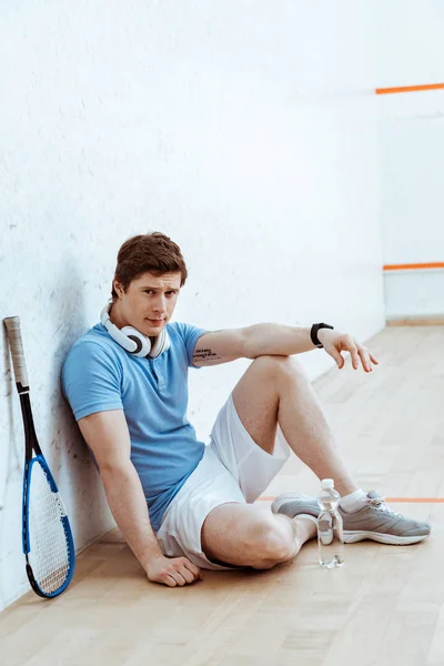 Triste joueur de squash en polo bleu assis sur le sol et regardant la caméra — Photo de stock