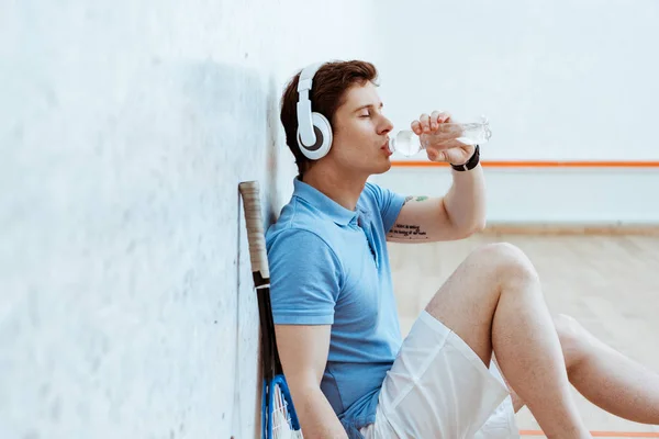 Squash player listening music in headphones and drinking water — Stock Photo