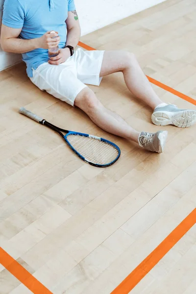 Vista recortada del jugador de squash sentado en el suelo y sosteniendo la botella de agua — Stock Photo