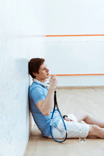 Jugador de squash cansado sentado en el suelo y sosteniendo la raqueta - foto de stock
