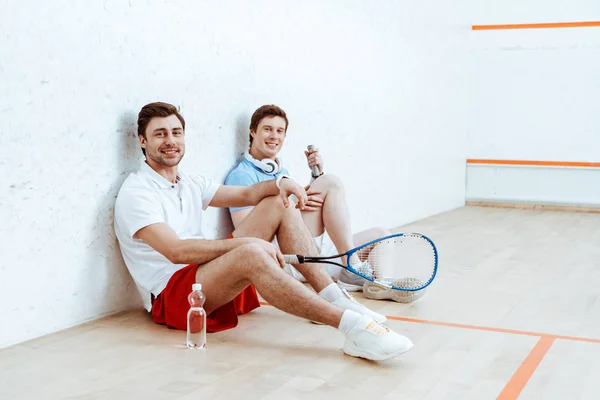 Два улыбающихся игрока в сквош сидят на полу в четырехстенном корте — стоковое фото
