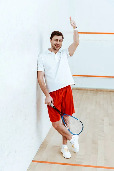 Vista completa del jugador de squash en pantalones cortos rojos sosteniendo la raqueta y mostrando el pulgar hacia arriba - foto de stock