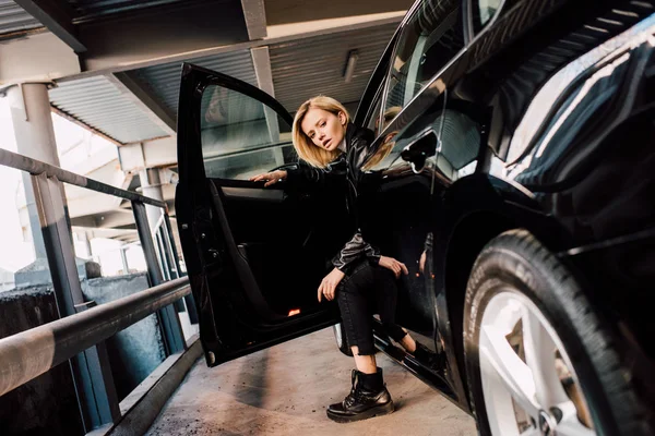 Attractive blonde girl sitting in car with open door and looking at camera — Stock Photo