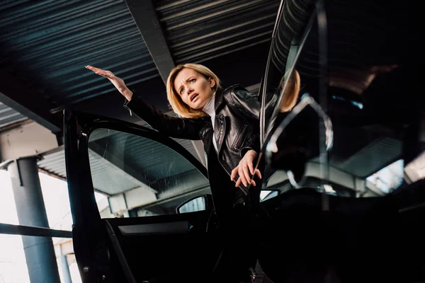 Low angle view of surprised blonde girl near black car with open door — Stock Photo