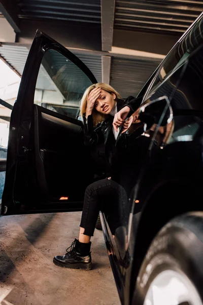 Emotional blonde woman holding head near black car with open door — Stock Photo
