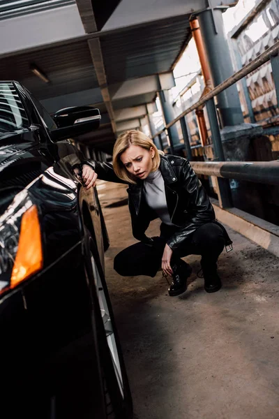 Verärgerte blonde Frau sitzt neben schwarzem Auto auf Parkplatz — Stockfoto