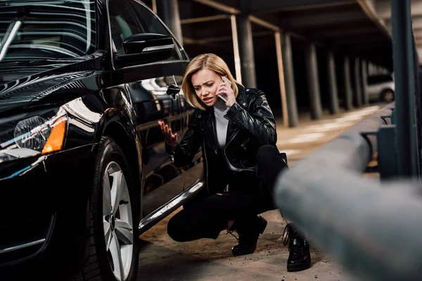 Verärgerte blonde Frau spricht auf Smartphone und gestikuliert, während sie neben Auto sitzt — Stockfoto