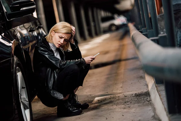 Расстроенная блондинка держит смартфон и трогает волосы, сидя рядом с машиной — стоковое фото