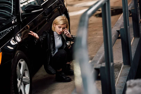 Selective focus of sad blonde girl talking on smartphone while sitting near black car — Stock Photo