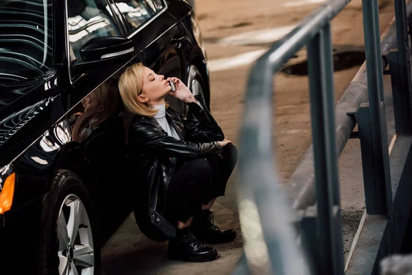 Вибірковий фокус привабливої блондинки, що говорить на смартфоні, сидячи біля чорного автомобіля — стокове фото