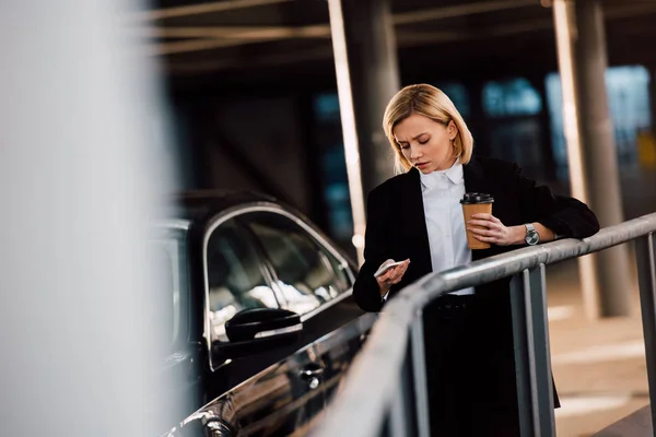 Foco seletivo de menina loira preocupada segurando smartphone e copo de papel perto de carro preto — Fotografia de Stock